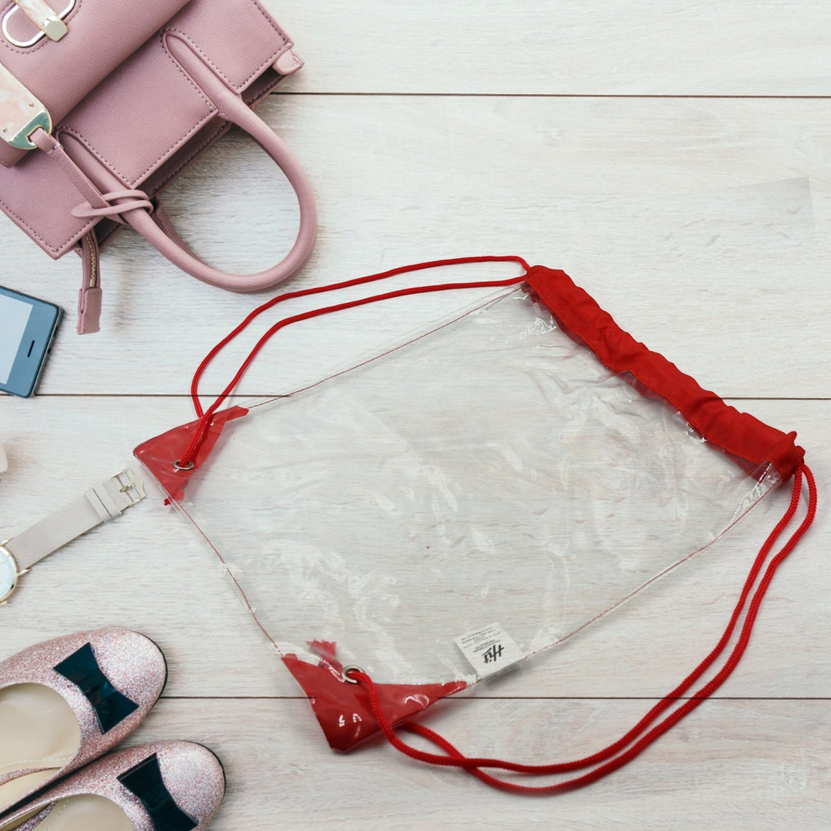 A clear ClassiCarry drawstring bag with red accents lies on a white wooden floor surrounded by a pink purse, phone, watch, Main image